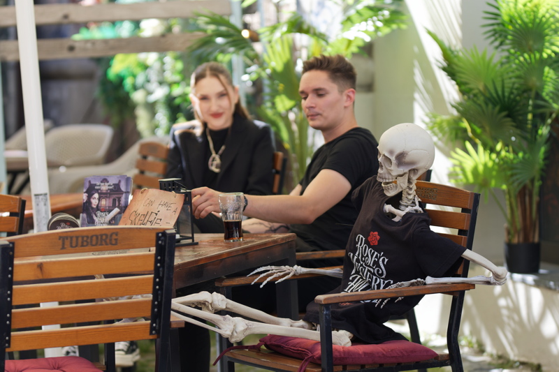 1 - A skeleton wearing a black graphic t-shirt sitting at a wooden café table